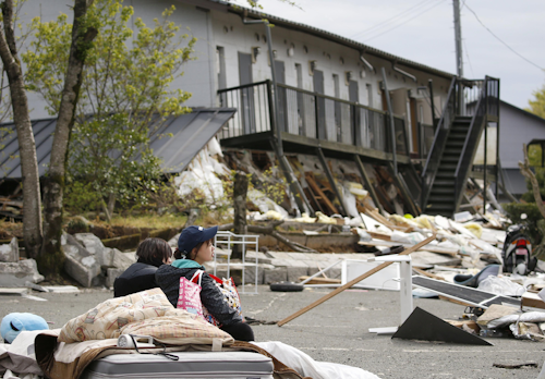 A Tokai University student, right, takes a rest after escaping from a collapsed student dormitory, background, following a powerful earthquake in Minamiaso village, Kumamoto prefecture, Japan, Saturday, April 16, 2016. The powerful earthquake struck southwestern Japan early Saturday, barely 24 hours after a smaller quake hit the same region. (Yuki Sato/Kyodo News via AP)