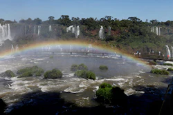 FILE - In this March 15, 2015 file photo, a tourist, far right, looks out at Iguazu Falls where a rainbow appears, seen from the Brazilian side in Foz do Iguazu, Brazil. Coca-Cola and Nestle say they aren’t negotiating for rights to the Guarani aquifer system, a massive fresh water reserve in South America, as online stories falsely claim. The stories incorrectly say the food and beverage companies are far along in talks to privatize for more than 100 years the underground reservoir, which provides water to people in Argentina, Brazil, Paraguay and Uruguay. Both companies issued statements saying the stories aren’t true. Iguazu Falls is located on Brazil's border with Argentina. (AP Photo/Jorge Saenz, File)