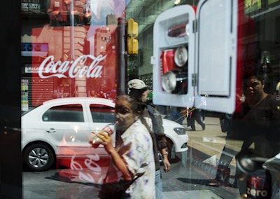 In this Oct. 9, 2014 file photo, a woman is reflected in a Coca-Cola store window display as she drinks a Coke in Mexico City. Image credit: AP Photo/Rebecca Blackwell, File