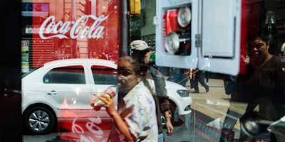 FILE - In this Oct. 9, 2014 file photo, a woman is reflected in a Coca-Cola store window display as she drinks a Coke in Mexico City. Mexicans are among the biggest soda drinkers in the world, so residents of the southern city of Ciudad Altamirano were hit hard when first Coca-Cola then Pepsi closed their distribution centers amid drug gang extortion demands. (AP Photo/Rebecca Blackwell, File)