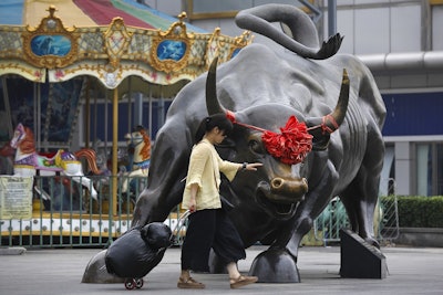 A woman pulls a 2 wheel trolley loaded with goods touches a bull statue on display outside a retail and wholesale clothing mall in Beijing, Monday, July 9, 2018. Image credit: AP Photo/Andy Wong