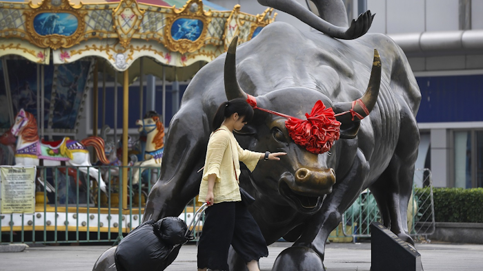 A woman pulls a 2 wheel trolley loaded with goods touches a bull statue on display outside a retail and wholesale clothing mall in Beijing, Monday, July 9, 2018. Image credit: AP Photo/Andy Wong