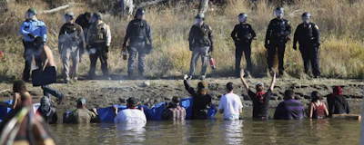 In this Nov. 2, 2016 file photo, protesters demonstrating against the expansion of the Dakota Access Pipeline wade in cold creek waters confronting local police as remnants of pepper spray waft over the crowd near Cannon Ball, N.D. North Dakota is demanding $38 million from the federal government to reimburse the state for costs associated with policing large-scale and prolonged protests against the oil pipeline. North Dakota Attorney General Wayne Stenehjem filed an administrative claim Friday, July 20, 2018. (AP Photo/John L. Mone)