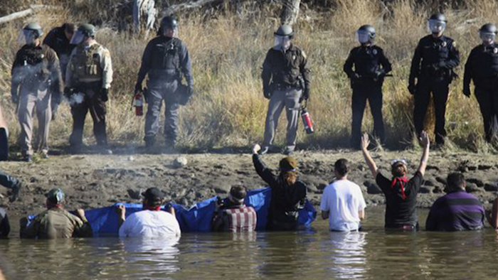 In this Nov. 2, 2016 file photo, protesters demonstrating against the expansion of the Dakota Access Pipeline wade in cold creek waters confronting local police as remnants of pepper spray waft over the crowd near Cannon Ball, N.D. North Dakota is demanding $38 million from the federal government to reimburse the state for costs associated with policing large-scale and prolonged protests against the oil pipeline. North Dakota Attorney General Wayne Stenehjem filed an administrative claim Friday, July 20, 2018. (AP Photo/John L. Mone)