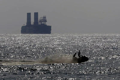 In this Monday, July 16, 2018 photo, two men ride a jet ski off the Lebanese coast as a floating power station waits off the coast at Jiyeh, south of Beirut, Lebanon. Lebanon received a floating power station from Turkey this week to ease the rolling blackouts that are a feature of its sticky summers. The 235-megawatt Esra Sultan, built and operated by the private Turkish company Karadeniz Energy, was billed by Energy and Water Minister Cesar Abi Khalil as a temporary but thrifty measure to bridge part of Lebanon's chronic electricity deficit. (AP Photo/Bilal Hussein)