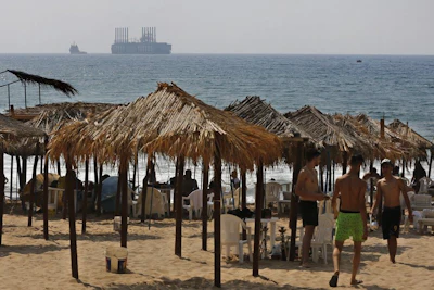 This Monday, July 16, 2018, photo shows people at the beach as a floating power station waits off the coast at Jiyeh, south of Beirut, Lebanon. Lebanon received a floating power station from Turkey this week to ease the rolling blackouts that are a feature of its sticky summers. The 235-megawatt Esra Sultan, built and operated by the private Turkish company Karadeniz Energy, was billed by Energy and Water Minister Cesar Abi Khalil as a temporary but thrifty measure to bridge part of Lebanon's chronic electricity deficit. (AP Photo/Bilal Hussein)