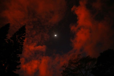 The moon is seen behind clouds glowing orange-red above a lava flow from a Kilauea volcano fissure on Hawaii's Big Island on May 19, 2018 in Kapoho, Hawaii. The U.S. Geological Survey said the volcano erupted explosively on May 17 launching a plume about 30,000 feet into the sky. (Photo by Mario Tama/Getty Images)
