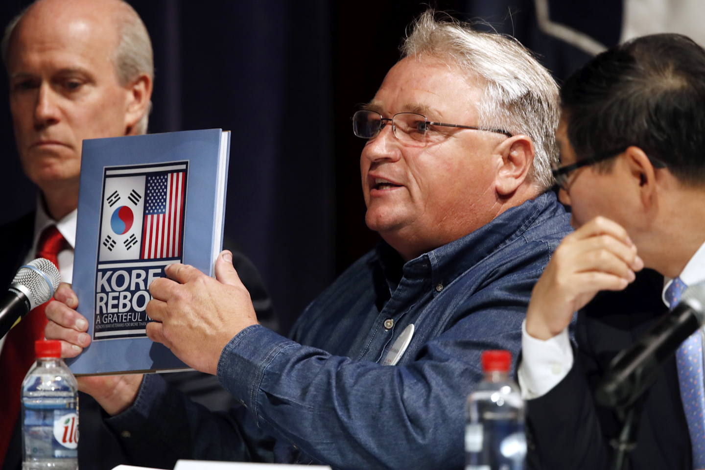 'We fought a war together,' says John Hall, a maintenance team member with Hyundai Motor Manufacturing Alabama, center, as he holds up a book about the Korean War, next to Joseph Boyle, senior director of business development and sales at LG Electronics, left, and Yong-Geun Kim, president and CEO of the Korean Automobile Manufacturers Association, during a Commerce Department hearing on investigation into whether auto imports threaten national security, Thursday, July 19, 2018, at the Commerce Department in Washington. The Commerce Department holds a daylong hearing on the latest trade fight the Trump administration is primed to start: It wants to impose tariffs on imported autos and auto parts, a move that President Donald Trump says would aid American workers but that could inflate car prices, make U.S. manufacturers less competitive and draw retaliation from other nations. (AP Photo/Jacquelyn Martin)