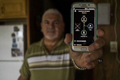 In this July 20 photo, Jose Santana poses for a picture showing his phone app where he can monitor the power generation and consumption of the solar system at his home in Las Piedras, Puerto Rico. Santana, a 64-year-old electronic technician, says he loves the smartphone app that lets him monitor the solar-charged Tesla batteries. He said the government should consider going solar and dumping the current 'archaic' power grid. (AP Photo/ Dennis M. Rivera Pichardo)