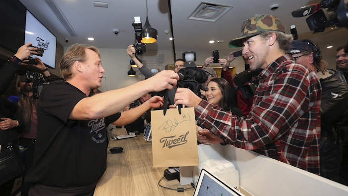 Canopy Growth CEO Bruce Linton, left to right, passes a bag with the first legal cannabis for recreation use sold in Canada to Nikki Rose and Ian Power at the Tweed shop on Water Street in St. John's N.L. at 12:01 am NDT on Wednesday Oct. 17, 2018. Image credit: Paul Daly/The Canadian Press via AP