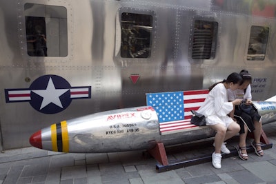 In this July 6, 2018, file photo, Chinese women look at phone near a rocket shaped bench with an American flag used as a marketing gimmick for a U.S. apparel shop in Beijing. Image credit: AP Photo/Ng Han Guan, File