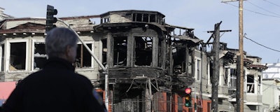 A San Francisco Police officer watches as a crew works at the scene of a Wednesday fire on Geary Boulevard in San Francisco, Thursday, Feb. 7, 2019. A gas explosion in shot a tower of flames into the sky and burned five buildings including one of the city's popular restaurants before firefighters brought the blaze under control. (AP Photo/Jeff Chiu)