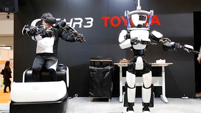 In this Dec. 18, 2019, photo, Toyota Motor Corp.'s human-shaped T-HR3 robot, right, is remotely controlled by its staff member, left, during a demonstration in Tokyo. Toyota's upgraded version of the human-shaped T-HR3 now has faster and smoother finger movements because the wearable remote-control device has become lighter and easier to use. The person wearing a headset and wiring made the robot move in exactly the same way he was moving, waving or making dance-like movements, as sensors sent computerized signals to the robot of what they detected as human movements.