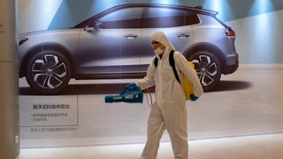In this Feb. 12, 2020, photo, a worker disinfects a mall near an advertisement for a car in Beijing, China. Automakers are gradually reopening factories in China that were idled by anti-virus control.