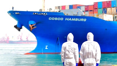 In this March 31, 2020 photo, workers in protective suits stand near a COSCO container ship docked at a port in Qingdao in eastern China's Shandong Province. China's government reported Tuesday, April 14, 2020 that exports fell further in March compared with February amid a global economic slowdown caused by the coronavirus pandemic.