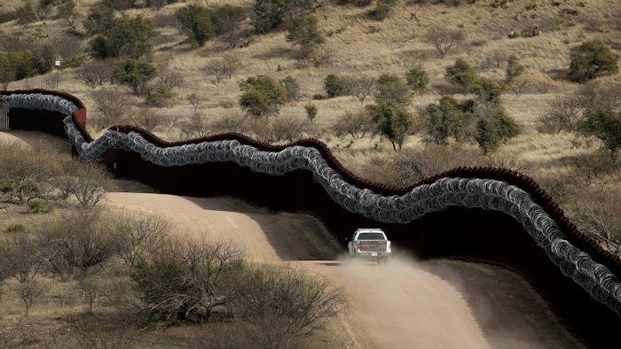 Customs and Border Control agent patrolling on the US side of a razor-wire-covered border wall.