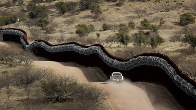 Customs and Border Control agent patrolling on the US side of a razor-wire-covered border wall.