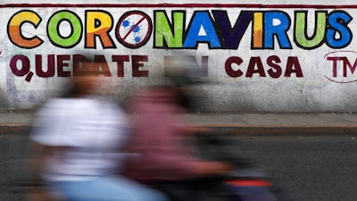 In this April 28, 2020 photo, a couple rides a motorcycle past a painted sign reading in Spanish 'Coronavirus. Stay Home,' in Iztapalapa, Mexico City. Mexico has had extremely limited testing compared with other countries, another factor that experts have said made it difficult for the country to contain the virus.