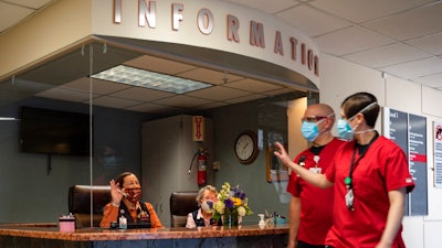 Medical Center Hospital auxiliary volunteers Gracie Sanchez, seated left, and Ann Ellison wave at passing health care workers as they work at Medical Center Hospital's information desk, Wednesday, March 10, 2021, in Odessa, Texas. In addition to wearing masks and remaining behind a plexiglass protective shield, the volunteers are required to have had a COVID-19 vaccine or be able to prove they have antibodies to the virus before returning to volunteer in person.