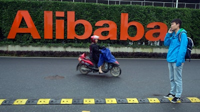 In this May 27, 2016, file photo, a man talks on his phone as a woman rides on an electric bike past a company logo at the Alibaba Group headquarters in Hangzhou in eastern China's Zhejiang province.