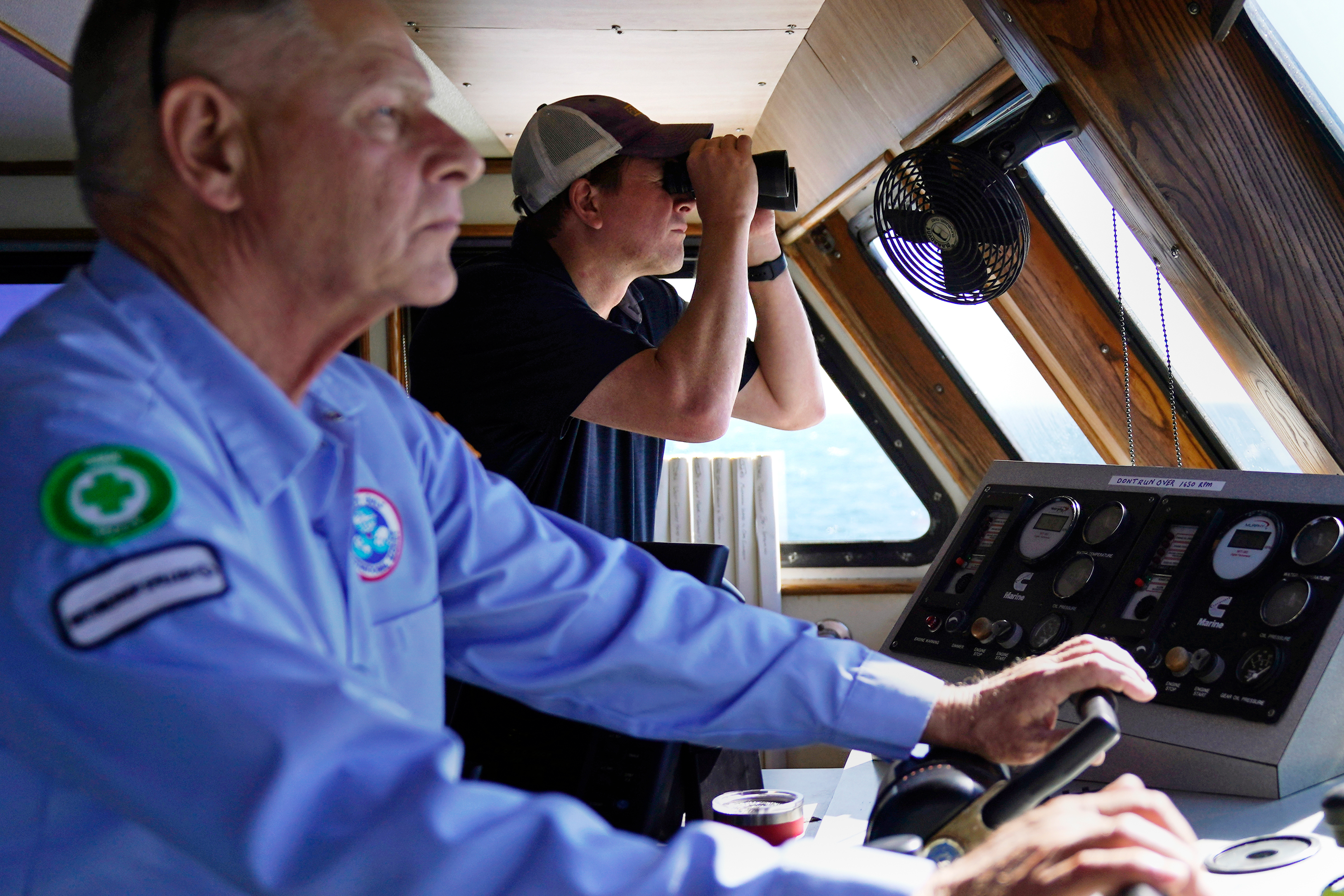 Aaron Smith, President and CEO of the Offshore Marine Service Association, center, peers through binoculars at ships installing the South Fork Wind project, as Capt. Rick Spaid, left, pilots the vessel Jones Act Enforcer, Tuesday, July 11, 2023, off the coast of Rhode Island.