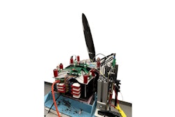 A battery sits on a wooden structure, hooked up to a propeller via exposed wires and electronic boards.