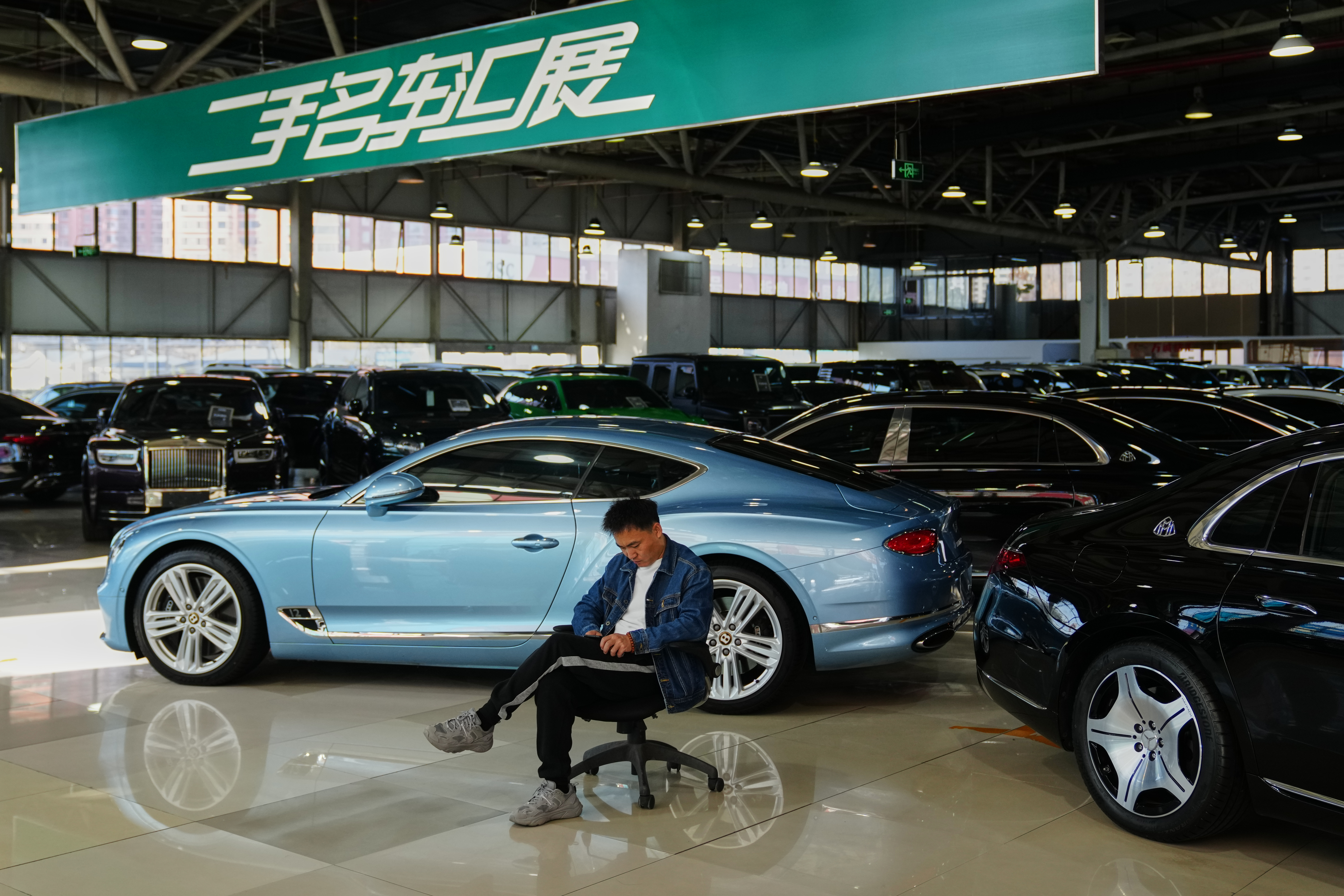 A man sits near luxury cars on offer at a second hand market in Beijing, Tuesday, Nov. 25, 2025.
