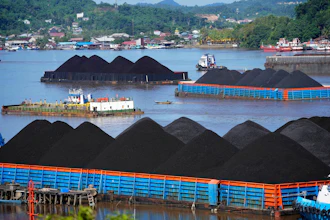 Industrial port with coal stockpiles, cargo barges, and vessels on blue water surrounded by forested mountains