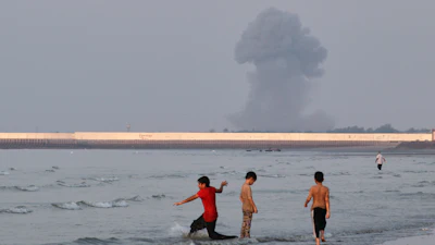 Three boys play in the shallow waters of the Strait of Hormuz as a plume of smoke rises from an explosion in the background off Bandar Abbas, Iran, July 13, 2026.
