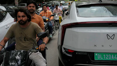 Two wheelers drive past an electric vehicle parked along a street in New Delhi, India, Monday, July 13, 2026.