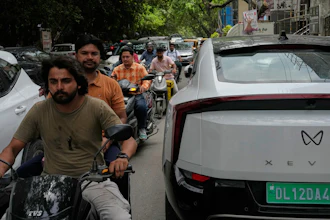 Two wheelers drive past an electric vehicle parked along a street in New Delhi, India, Monday, July 13, 2026.