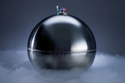 Polished silver spherical object with ornamental finial surrounded by dry ice vapor on dark background