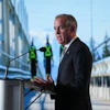 Professional man speaking at podium during outdoor event with construction workers and modern buildings in background