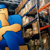 Cartoon warehouse worker in blue uniform sitting in front of industrial shelving with stacked boxes