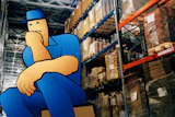 Cartoon warehouse worker in blue uniform sitting in front of industrial shelving with stacked boxes