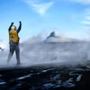 Person in yellow vest stands triumphantly on volcanic landscape with UFO and spacecraft visible in cloudy sky
