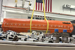Orange cylindrical aerospace equipment suspended by yellow straps in industrial workshop with American flags