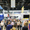 Crowded trade show exhibition floor with multiple vendor booths, signage, and professional attendees networking in an indoor convention center.
