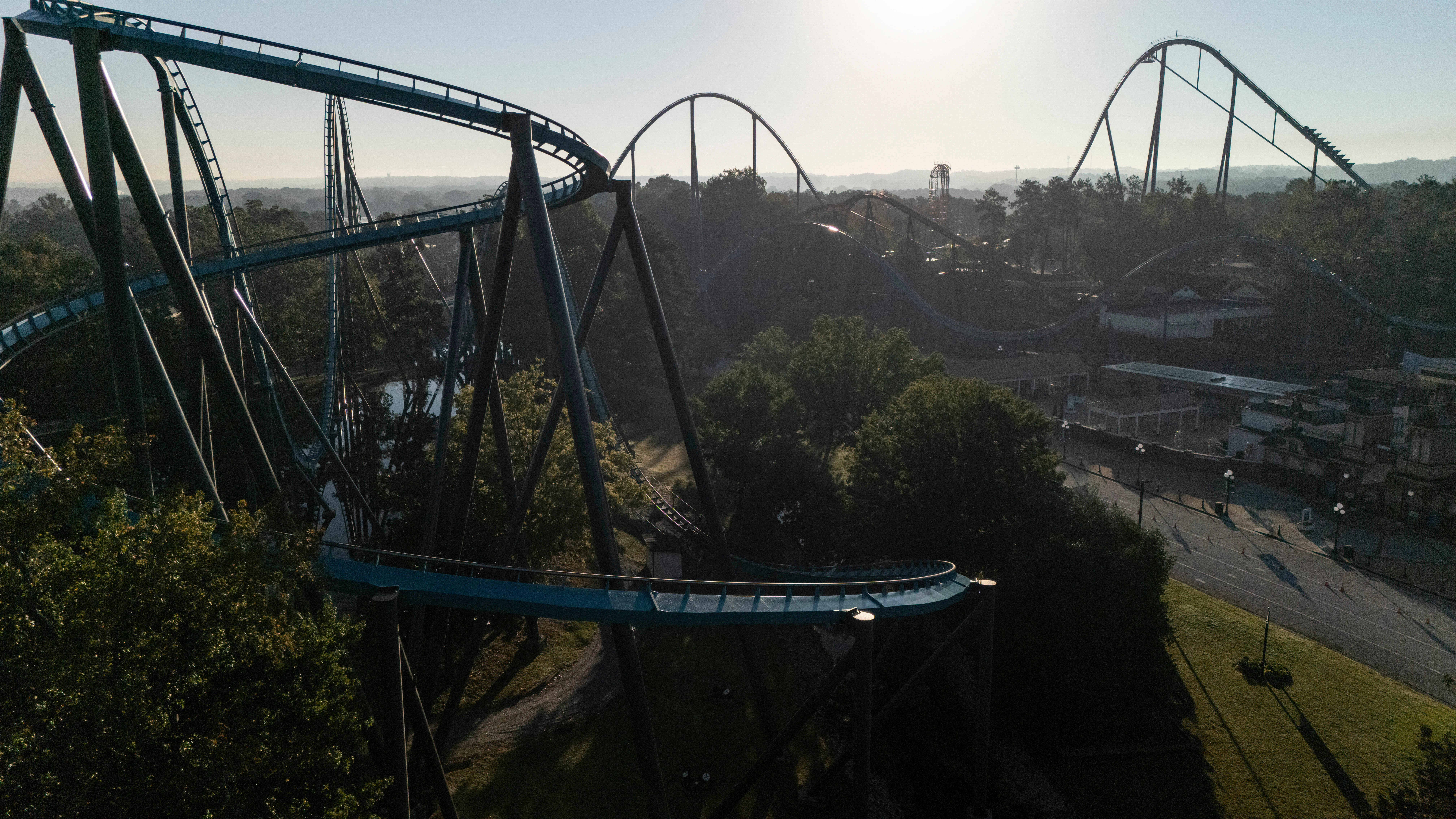 Aerial view of blue roller coaster tracks winding through an amusement park surrounded by trees and landscape