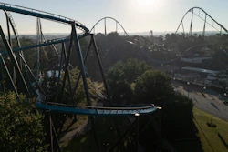 Aerial view of blue roller coaster tracks winding through an amusement park surrounded by trees and landscape