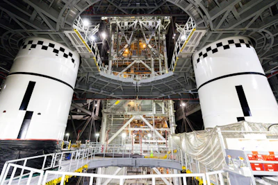 Two white spacecraft modules with black checkered docking targets in assembly facility with metal framework and equipment overhead.