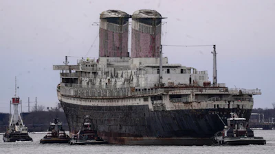 Large cargo ship assisted by tugboats in port waters with industrial twin smokestacks visible