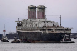 Large cargo ship assisted by tugboats in port waters with industrial twin smokestacks visible