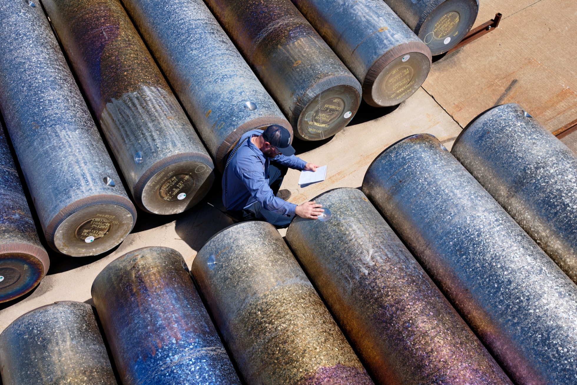 Worker in blue surveying large industrial metal pipes arranged on sandy ground from above