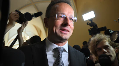 Man in suit and glasses at press conference surrounded by media cameras and microphones