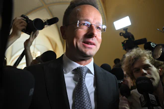 Man in suit and glasses at press conference surrounded by media cameras and microphones