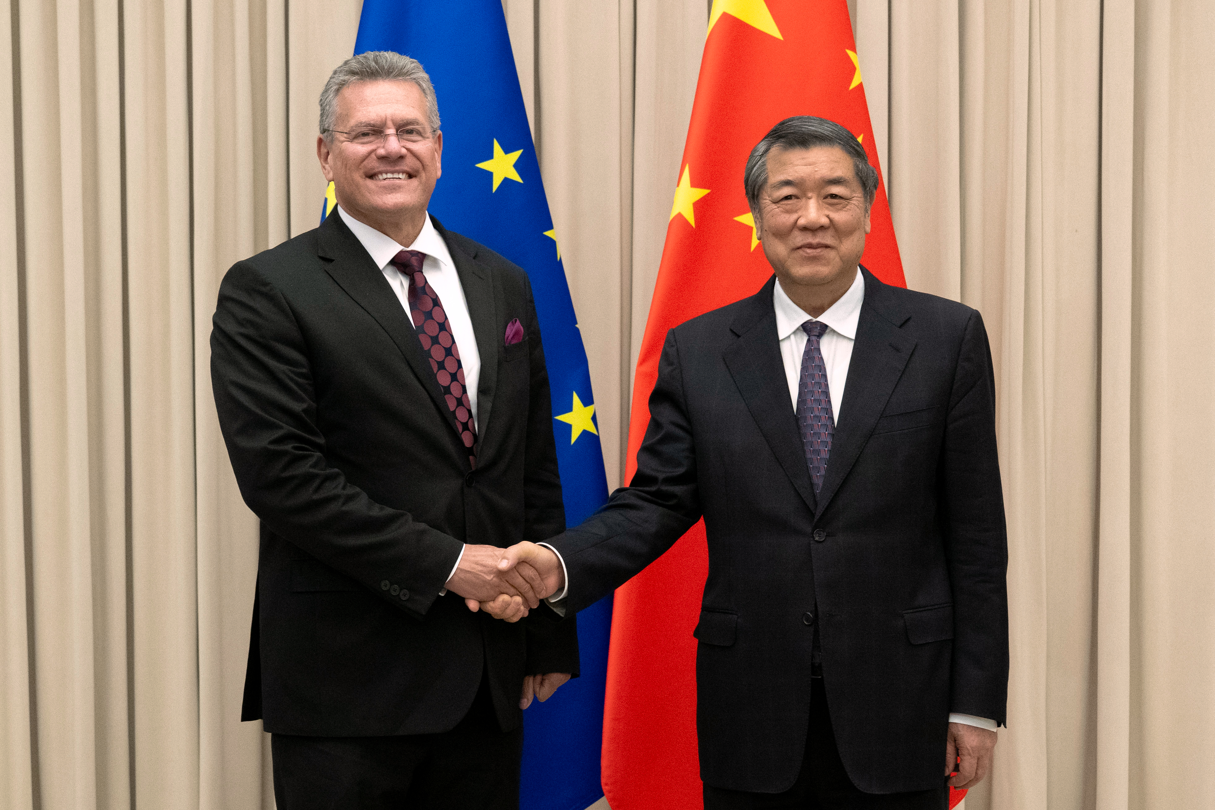 Two officials in formal business attire shake hands at a diplomatic meeting with EU and Chinese flags in the background.