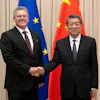 Two officials in formal business attire shake hands at a diplomatic meeting with EU and Chinese flags in the background.