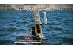 Autonomous solar-powered sailboat with white sails on blue water near waterfront buildings