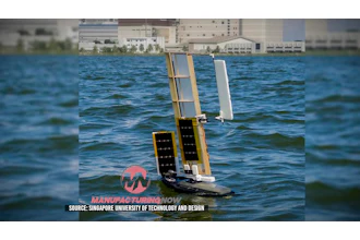 Autonomous solar-powered sailboat with white sails on blue water near waterfront buildings